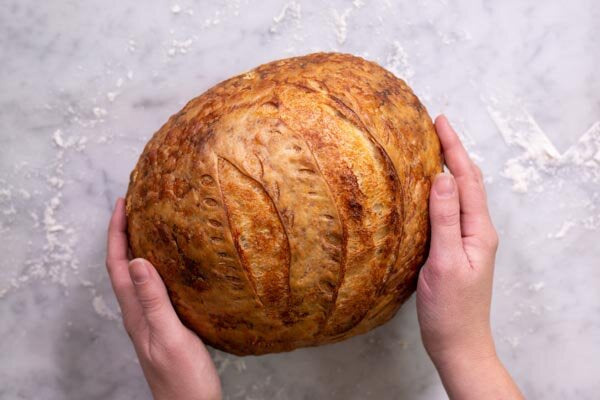 Hands cradling a golden scored sourdough boule on a marble surface