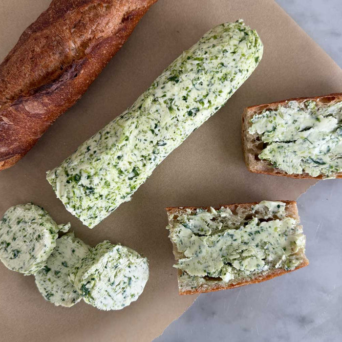 A log of bright green ramp butter on parchment paper, with three thick coins sliced off, a torn baguette, and two bread slices already spread with ramp butter on a marble surface.
