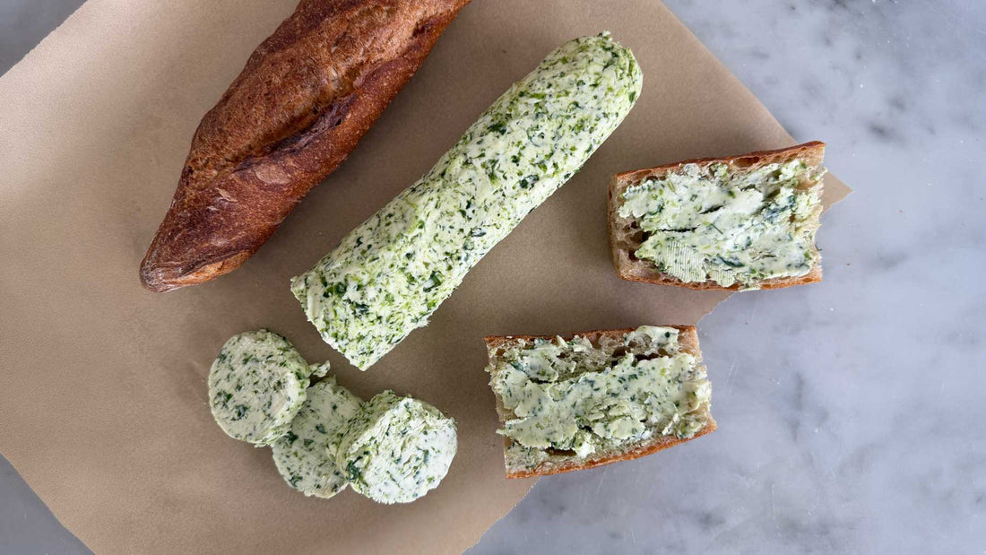 A log of bright green ramp butter on parchment paper, with three thick coins sliced off, a torn baguette, and two bread slices already spread with ramp butter on a marble surface.