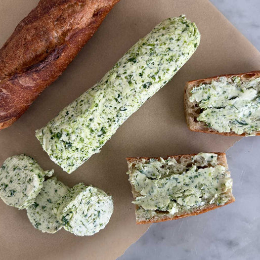 A log of bright green ramp butter on parchment paper, with three thick coins sliced off, a torn baguette, and two bread slices already spread with ramp butter on a marble surface.