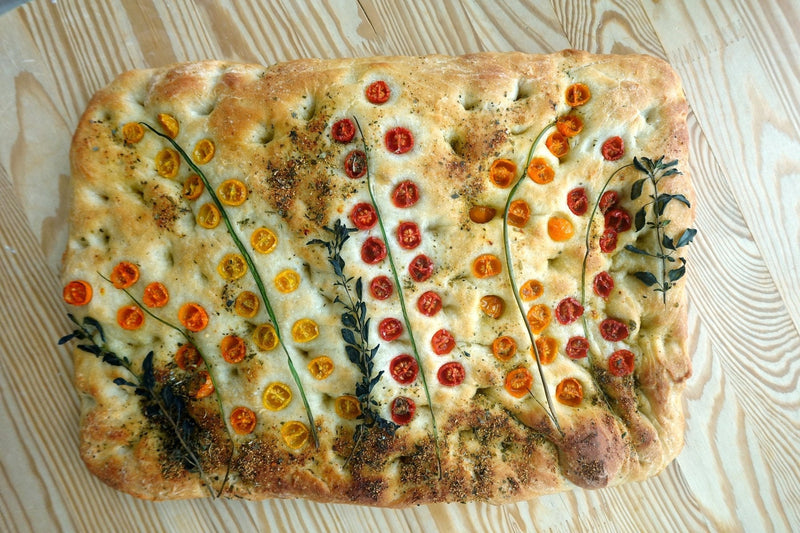 Garden focaccia decorated with rows of halved cherry tomatoes and fresh herb sprigs, seen from above
