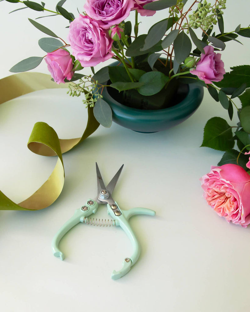 Lagoon green pruning shears beside a vase of pink roses and eucalyptus