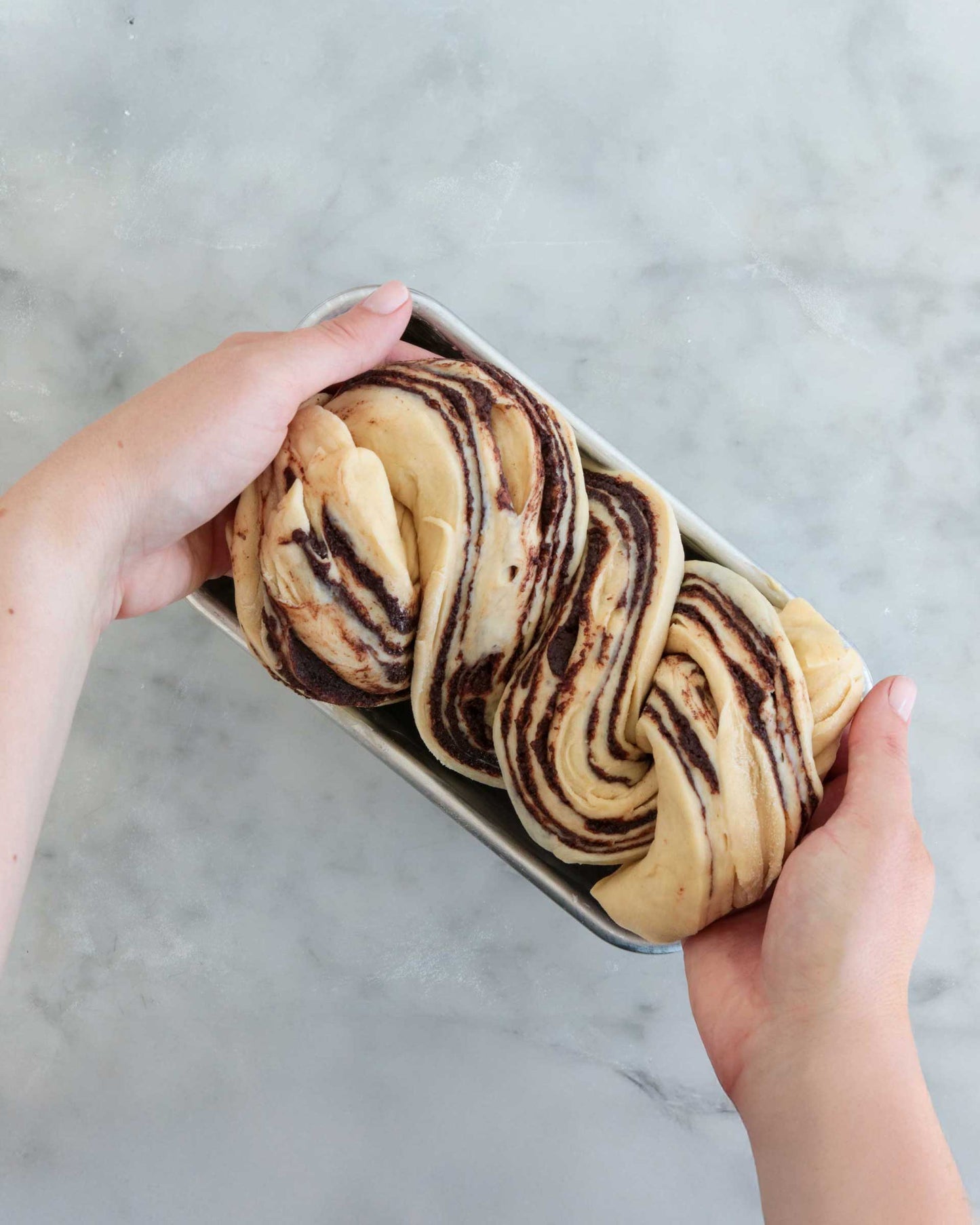 Hands placing twisted chocolate babka dough into a loaf pan