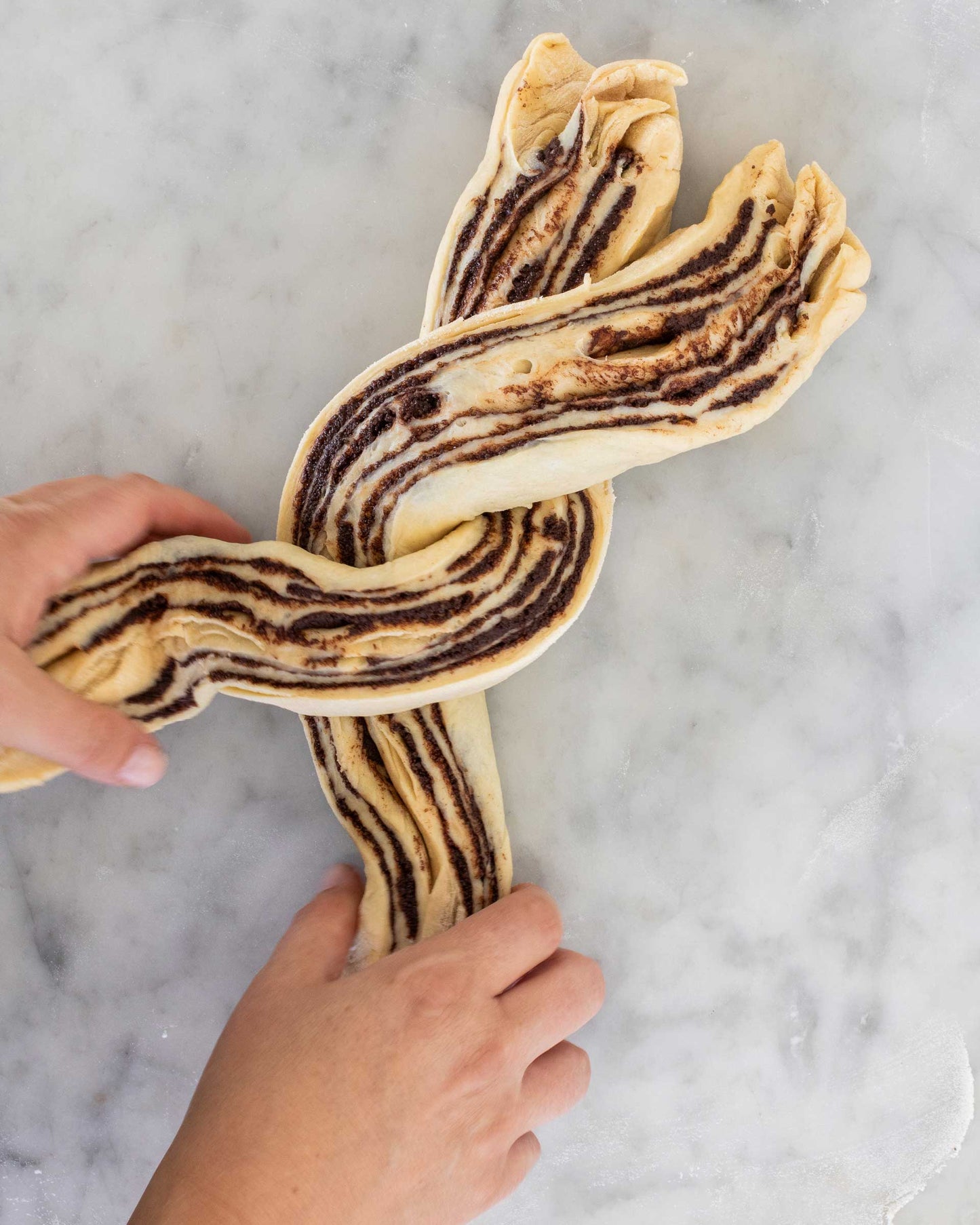 Hands twisting chocolate babka dough on a marble counter