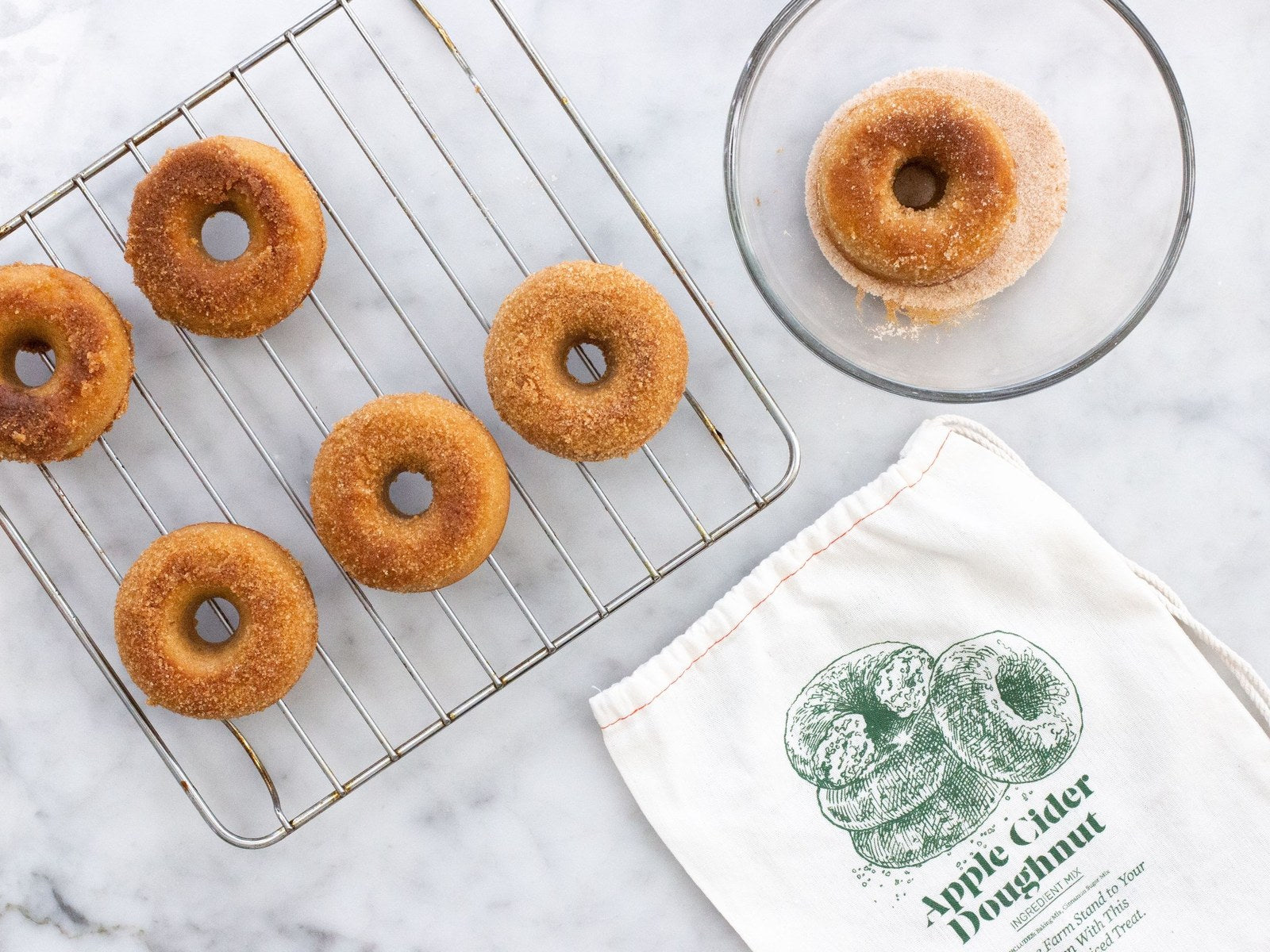Apple cider doughnuts on a cooling rack with the FarmSteady mix bag and a bowl of cinnamon sugar