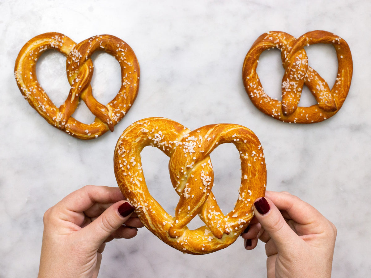 Hands holding a salted soft pretzel made with the FarmSteady Soft Pretzel Baking Mix, two more on marble